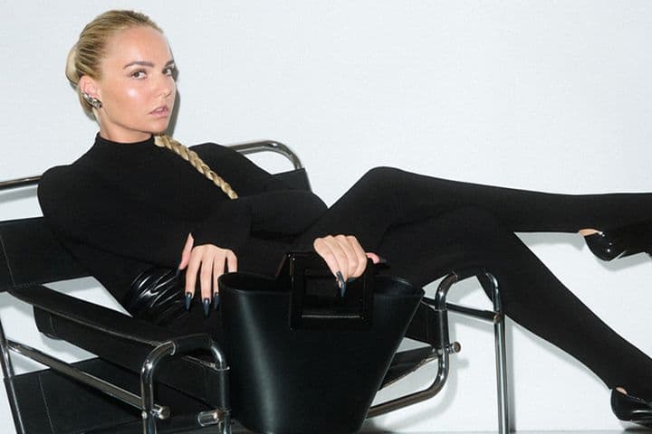 Γυναίκα με μακριά ξανθή κοτσίδα, φοράει εφαρμοστό μαύρο σύνολο και γόβες με μυτερή μύτη, είναι ξαπλωμένη σε μοντέρνα μαύρη μεταλλική καρέκλα και κρατά δομημένη μαύρη τσάντα, με λευκό φόντο.