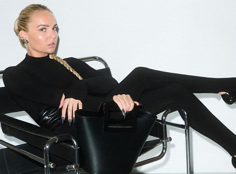 Γυναίκα με μακριά ξανθή κοτσίδα, φοράει εφαρμοστό μαύρο σύνολο και γόβες με μυτερή μύτη, είναι ξαπλωμένη σε μοντέρνα μαύρη μεταλλική καρέκλα και κρατά δομημένη μαύρη τσάντα, με λευκό φόντο.