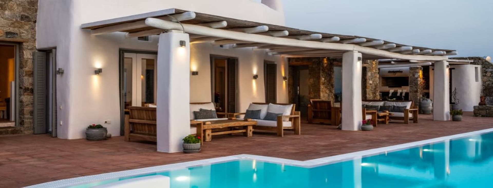 An outdoor Mediterranean-style terrace with a wooden pergola, white columns, wooden lounge furniture, terracotta floor tiles, and a swimming pool.