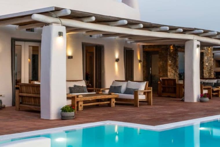 An outdoor Mediterranean-style terrace with a wooden pergola, white columns, wooden lounge furniture, terracotta floor tiles, and a swimming pool.