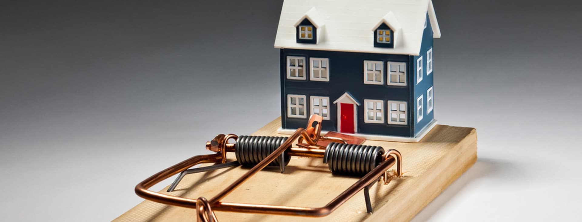 Ein kleines Miniaturhaus ist auf einer großen Mausefalle platziert, was die potenziellen Risiken im Zusammenhang mit dem Besitz eines Hauses oder dem Immobilienmarkt symbolisiert.