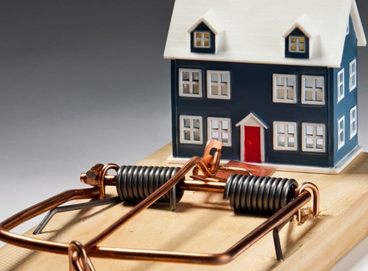 Ein kleines Miniaturhaus ist auf einer großen Mausefalle platziert, was die potenziellen Risiken im Zusammenhang mit dem Besitz eines Hauses oder dem Immobilienmarkt symbolisiert.