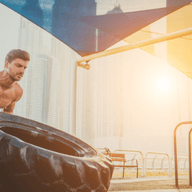 Man lifting a large tire outdoors at a gym with city buildings in the background, under a canopy with sunlight streaming through.
