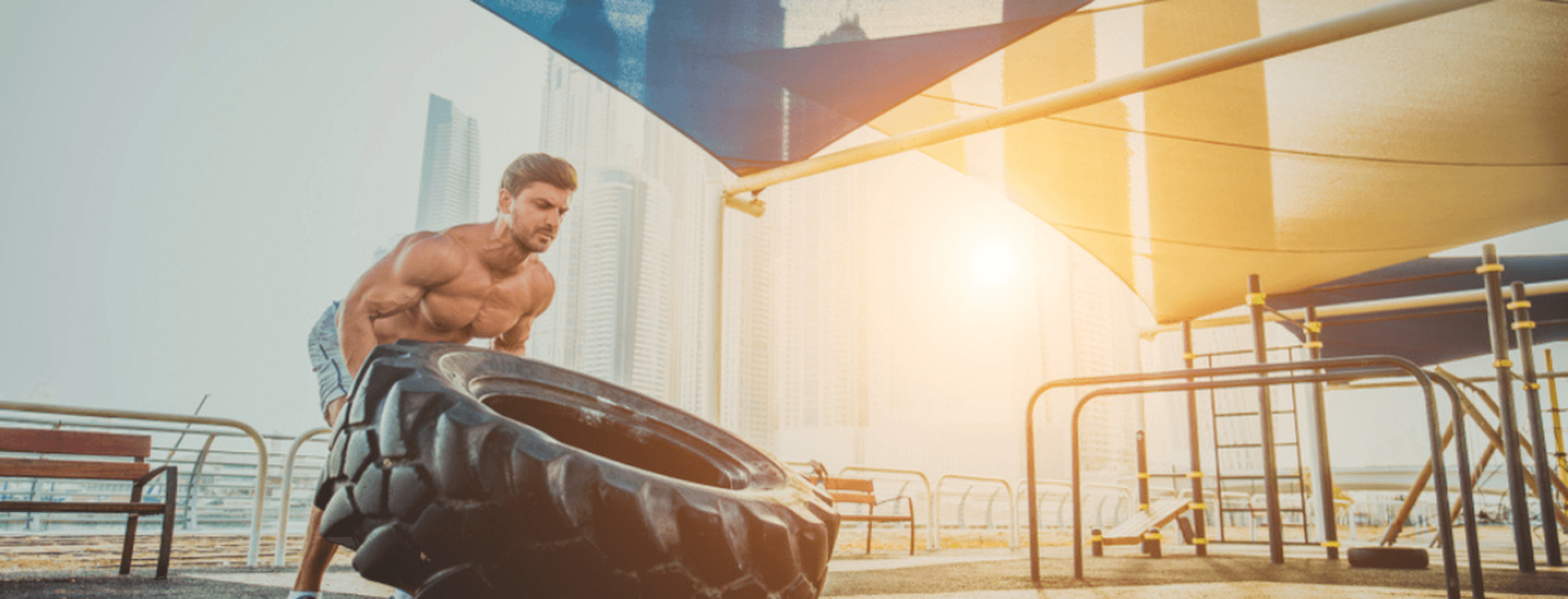 Man lifting a large tire outdoors at a gym with city buildings in the background, under a canopy with sunlight streaming through.