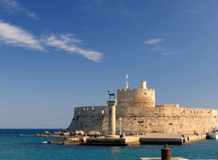 Harbor view of Rhodes with the medieval fortress and the iconic deer statue on a column overlooking the sea under a clear blue sky.