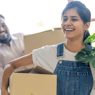 A smiling woman carries a box and a potted plant, followed by a man with another box, as they move into a bright room.