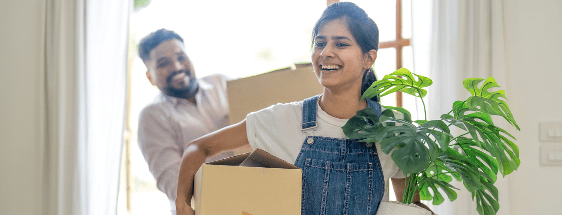 A smiling woman carries a box and a potted plant, followed by a man with another box, as they move into a bright room.