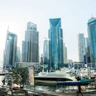 Panoramic view of a marina with luxury yachts and high-rise buildings under a clear sky, surrounded by a boardwalk and greenery.