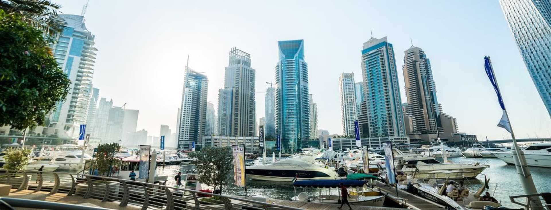 Panoramic view of a marina with luxury yachts and high-rise buildings under a clear sky, surrounded by a boardwalk and greenery.