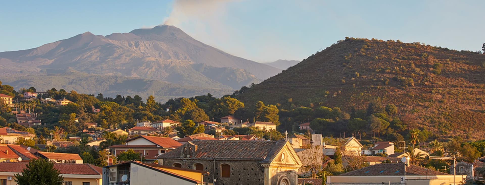 Ein malerischer Blick auf eine Stadt mit Häusern und einer Kirche vor dem Hintergrund eines rauchenden Vulkans und Hügeln unter einem klaren Himmel.