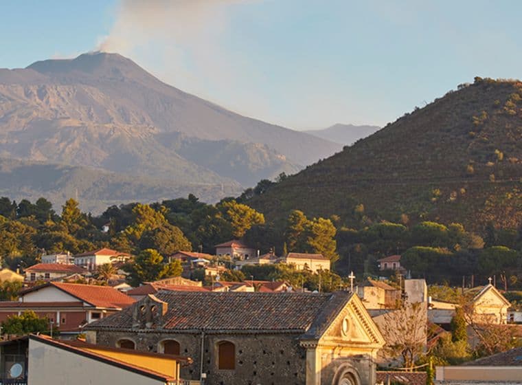 Ein malerischer Blick auf eine Stadt mit Häusern und einer Kirche vor dem Hintergrund eines rauchenden Vulkans und Hügeln unter einem klaren Himmel.