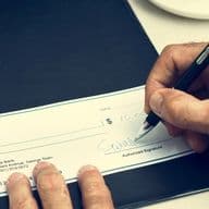 Person signing a check with a pen on a black folder, wearing a blue checkered shirt.