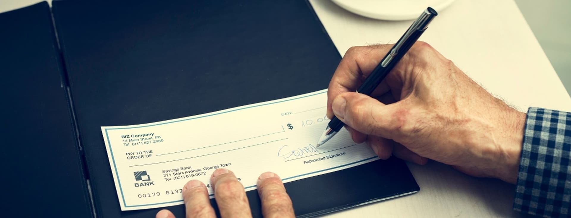 Person signing a check with a pen on a black folder, wearing a blue checkered shirt.