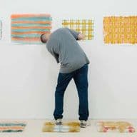 A person looks at colourful abstract artworks on a white wall in a gallery, with similar artworks displayed on the floor.