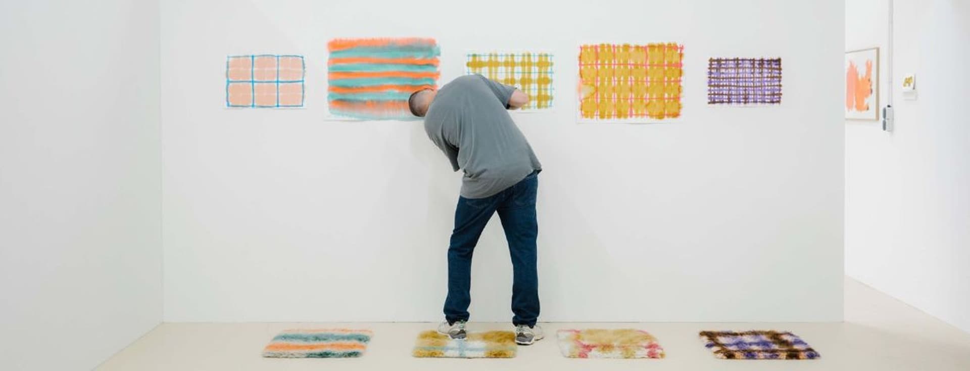 Eine Person betrachtet farbenfrohe abstrakte Kunstwerke an einer weißen Wand in einer Galerie, wobei ähnliche Kunstwerke auf dem Boden ausgestellt sind.