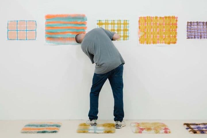 A person looks at colourful abstract artworks on a white wall in a gallery, with similar artworks displayed on the floor.