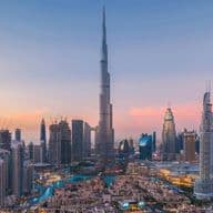 Panoramic view of the Dubai skyline at sunset, featuring a prominent tall skyscraper surrounded by various modern buildings under a colorful sky.
