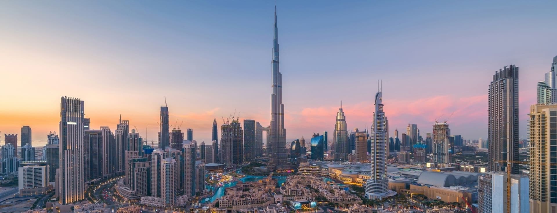 Die Skyline von Dubai bei Sonnenuntergang. Hohe Wolkenkratzer, darunter der Burj Khalifa, dominieren das Stadtbild. Der Himmel ist eine Mischung aus Orange, Rosa und Blau.