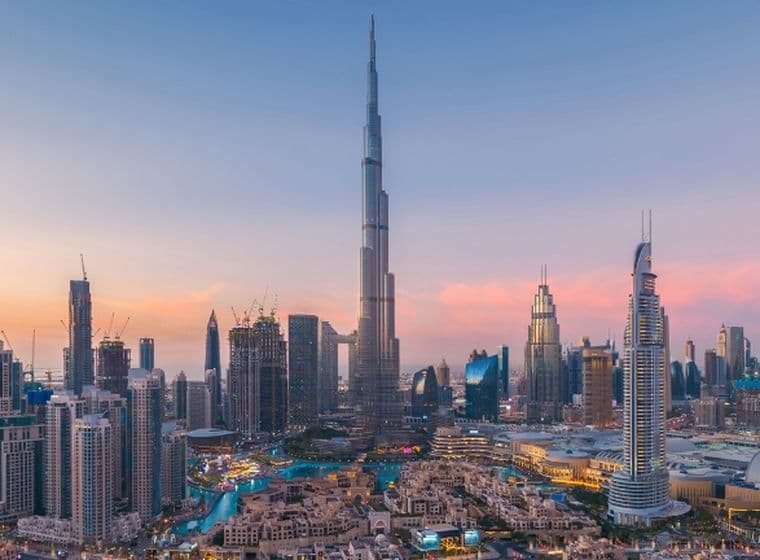 Panoramic view of the Dubai skyline at sunset, featuring a prominent tall skyscraper surrounded by various modern buildings under a colorful sky.