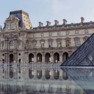 Die historische Fassade des Louvre und die moderne Glaspyramide spiegeln sich in einer ruhigen Wasserfläche unter einem klaren Himmel.