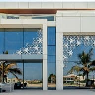 Modern luxury villa with large glass windows, intricate white lattice designs, and palm trees in the foreground. Outdoor seating area visible.