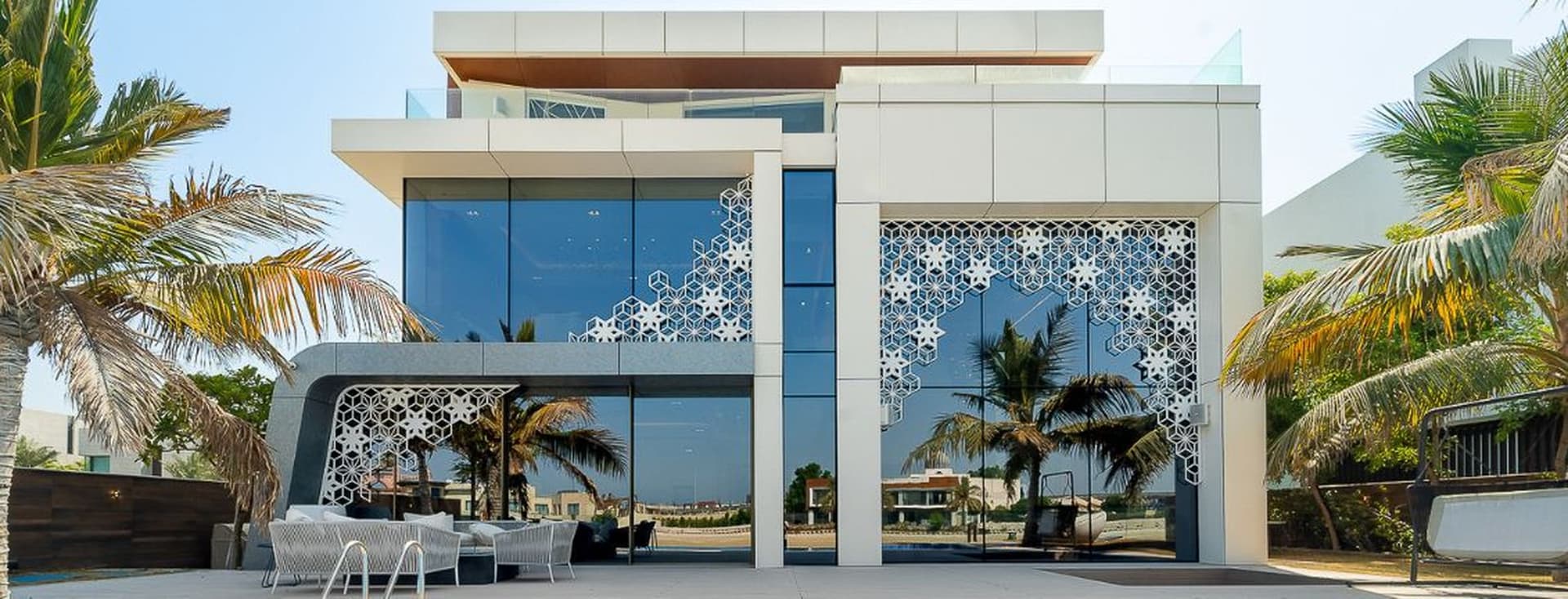 Modern luxury villa with large glass windows, intricate white lattice designs, and palm trees in the foreground. Outdoor seating area visible.