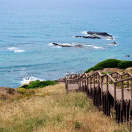 Wooden boardwalk descending a grassy hillside towards a rocky coastline with a vast, calm ocean under a clear sky.