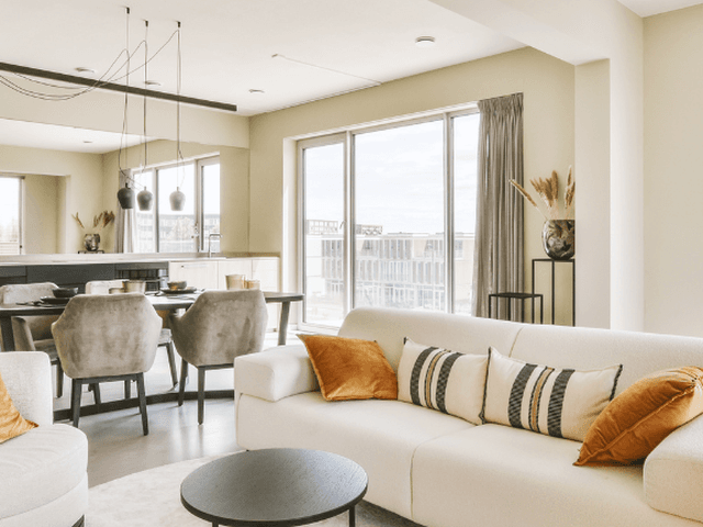 Helles, modernes Wohnzimmer mit einem weißen Sofa, orangefarbenen Kissen, Esstisch und großen Fenstern.