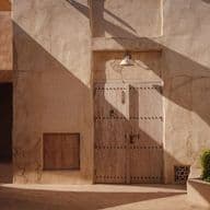 Außenansicht eines beigefarbenen Gebäudes mit einer Holztür, einem Torbogen und grünen Büschen. Sonnenlicht wirft Schatten auf die strukturierten Wände.