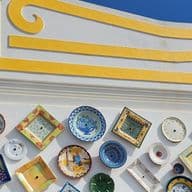 Eine Wand, geschmückt mit bunten, dekorativen Tellern in verschiedenen Formen und Mustern, vor einem weiß und gelb bemalten Hintergrund unter einem blauen Himmel.