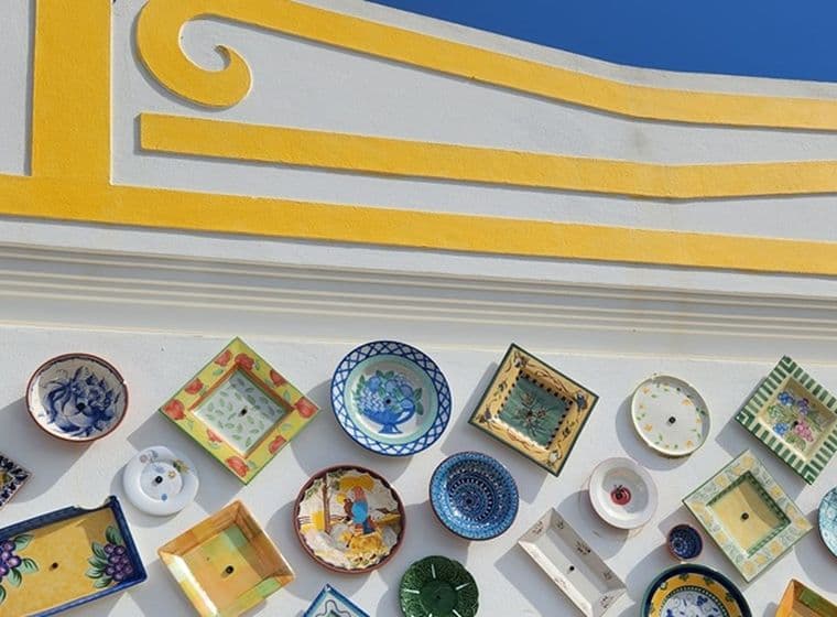 Eine Wand, geschmückt mit bunten, dekorativen Tellern in verschiedenen Formen und Mustern, vor einem weiß und gelb bemalten Hintergrund unter einem blauen Himmel.