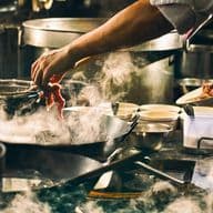 Chef adding ingredients to a steaming pan in a busy kitchen, surrounded by pots and bowls, with warm, ambient lighting.