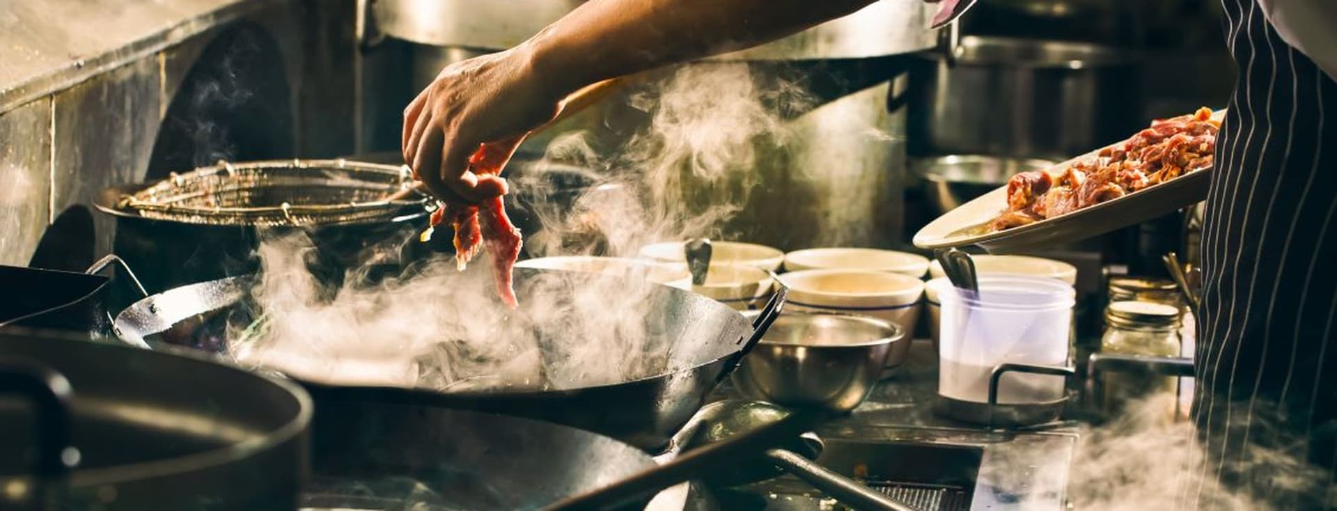 Chef adding ingredients to a steaming pan in a busy kitchen, surrounded by pots and bowls, with warm, ambient lighting.