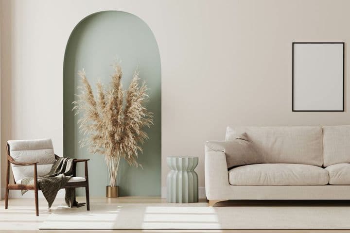 Minimalistisches Wohnzimmer mit einem beigen Sofa, einem Holzstuhl mit einer Decke, einer Vase mit Pampasgras in einer Nische und einem leeren Bilderrahmen.