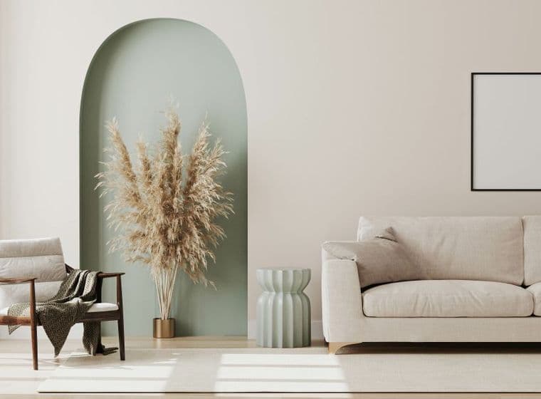 Minimalist living room with a beige sofa, a wooden chair with a blanket, a vase of pampas grass in an alcove, and an empty picture frame.