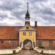 Historisches Backsteingebäude mit einem zentralen Torbogen, einer gelben Fassade und einem Uhrenturm. Bedeckter Himmel im Hintergrund.