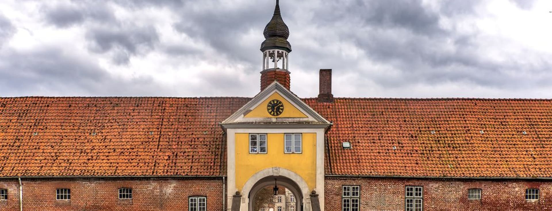 Historic brick building with a central archway, yellow facade, and a clock tower. Overcast sky in the background.