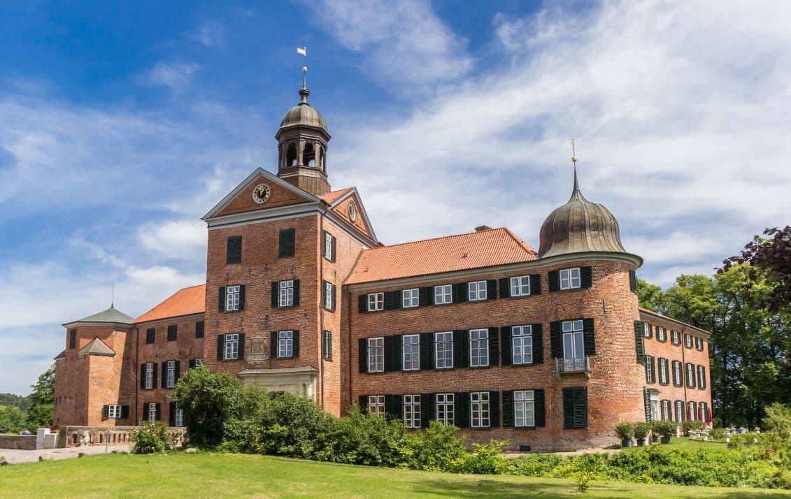 Stockelsdorf Manor house, a historic brick castle with a red-tiled roof, surrounded by greenery, under a blue sky with scattered clouds.