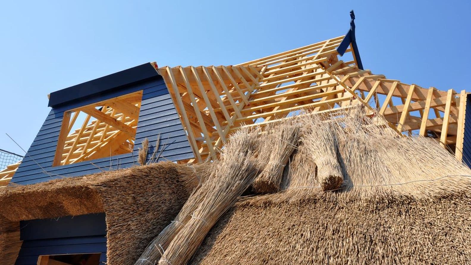 Thatched roof under construction with wooden framework and bundles of straw against a clear blue sky.