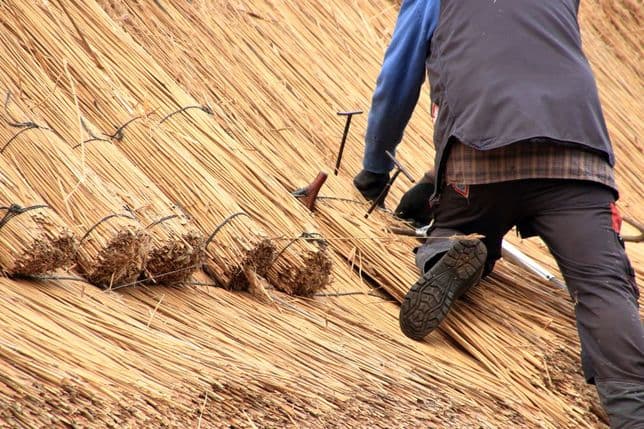 Person kneeling and working on a thatched roof, securing bundles of straw with tools.
