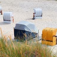 Strandszene mit mehreren gestreiften und einfarbigen Strandkörben am Sandstrand, umgeben von hohem Gras im Vordergrund.