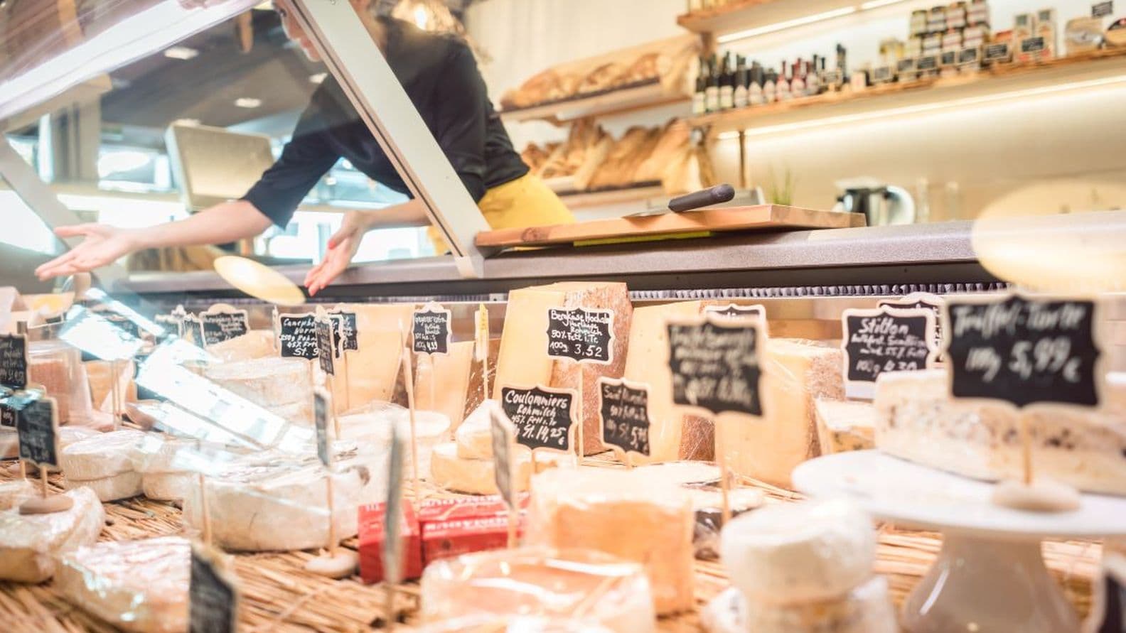 Cheese shop display with various types of cheeses labeled with prices on a wicker counter, shelves with bread and bottles in the background.
