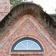 A brick gable with a semi-circular window is topped by a thatched roof, surrounded by trees in the background.