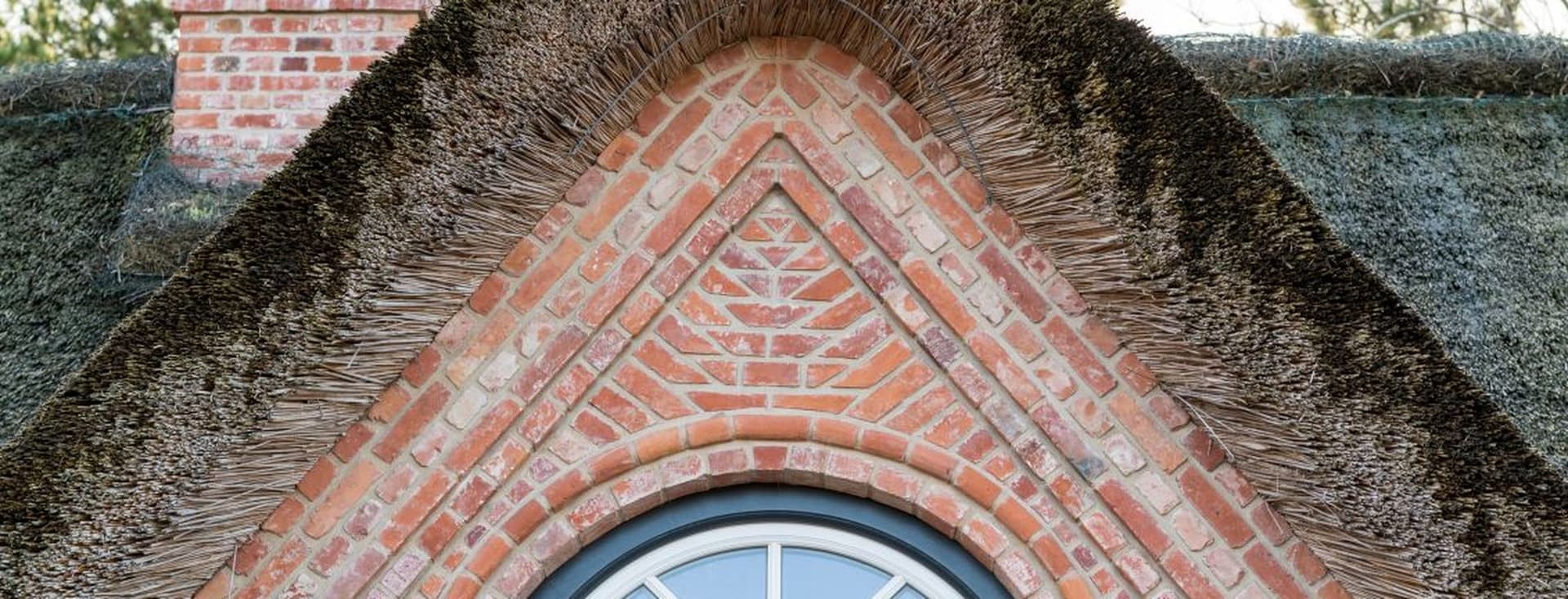 A brick gable with a semi-circular window is topped by a thatched roof, surrounded by trees in the background.