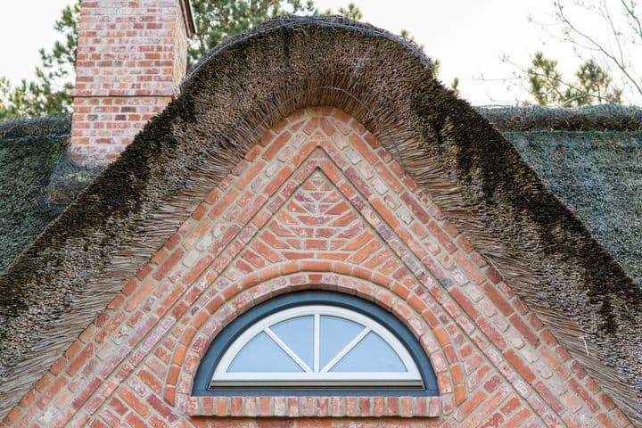Ein gemauerter Giebel mit einem halbrunden Fenster wird von einem Strohdach gekrönt, das im Hintergrund von Bäumen umgeben ist.