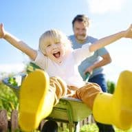 Ein fröhliches Kind mit ausgestreckten Armen sitzt in einer Schubkarre, die von einem Erwachsenen in einem sonnigen Garten geschoben wird, der gelbe Stiefel trägt.