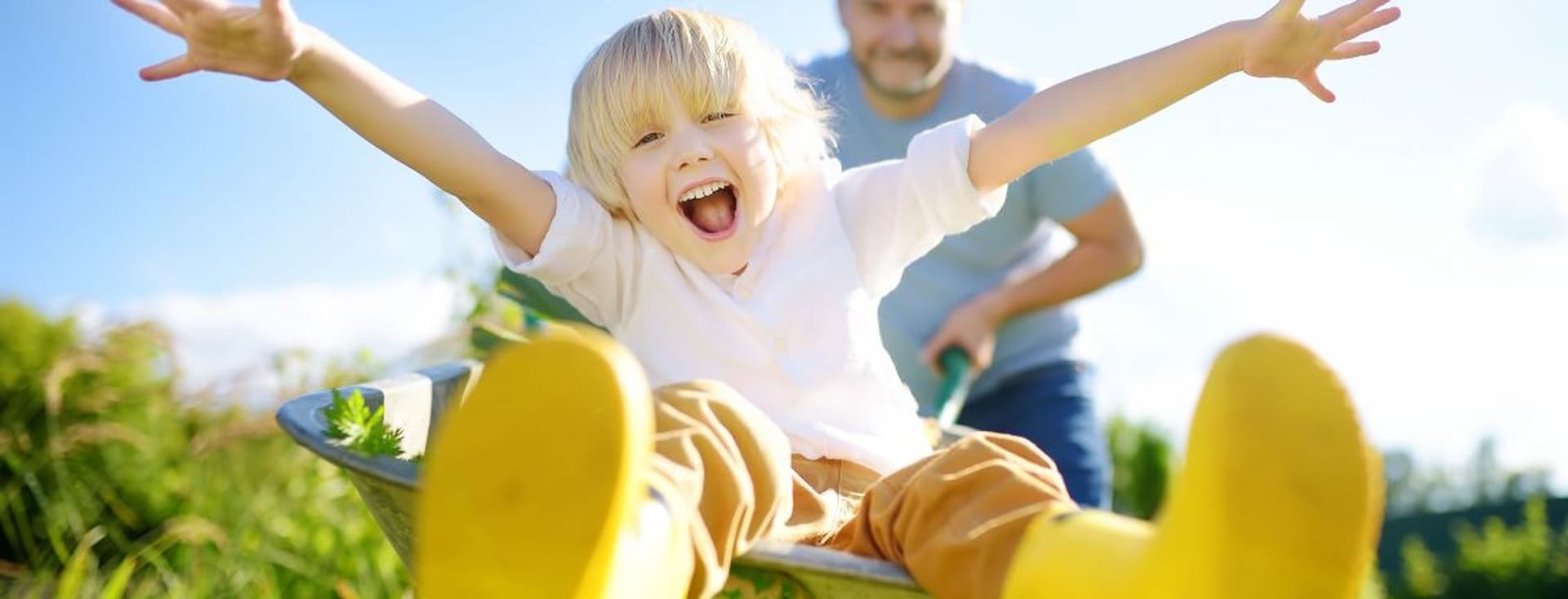 A joyful child with outstretched arms sits in a wheelbarrow, pushed by an adult in a sunny garden, wearing yellow boots.
