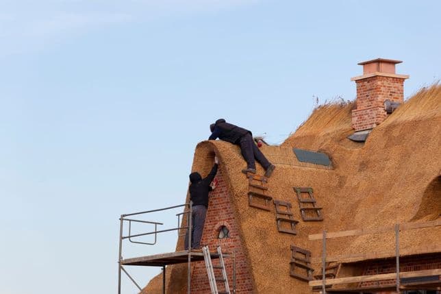 Two workers on scaffolding install a thatched roof on a brick house under a clear blue sky.