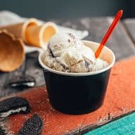 Ein Becher mit Keksen und Sahneeis mit einem roten Löffel auf einer rustikalen Holzfläche, umgeben von zerbrochenen Waffeltüten und Keksen.