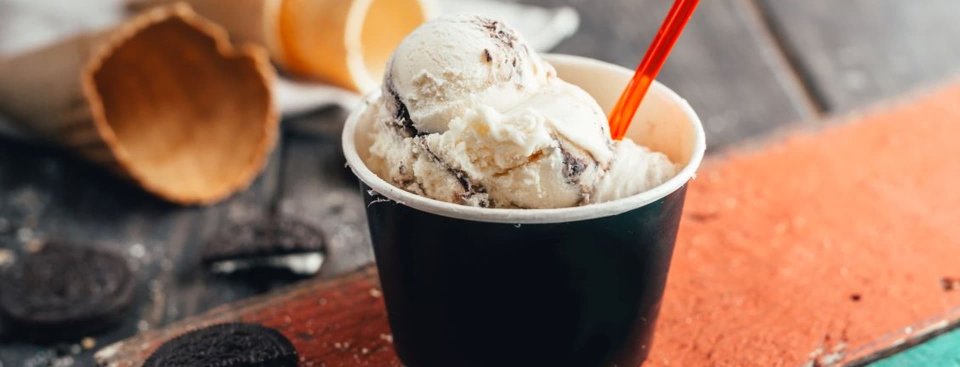 A cup of cookies and cream ice cream with a red spoon on a rustic wooden surface, surrounded by broken waffle cones and cookies.